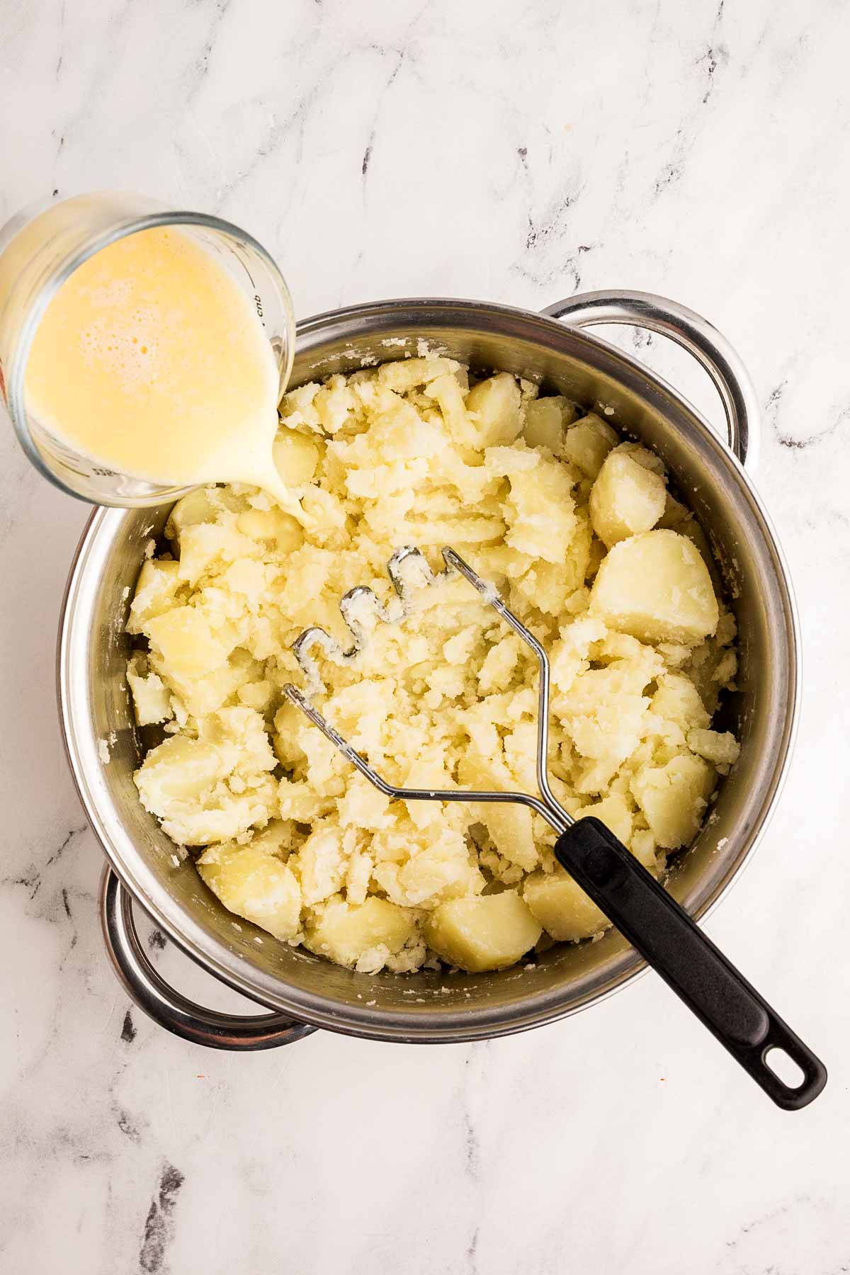 milk pouring from glass measuring jug into pot of mashed potatoes